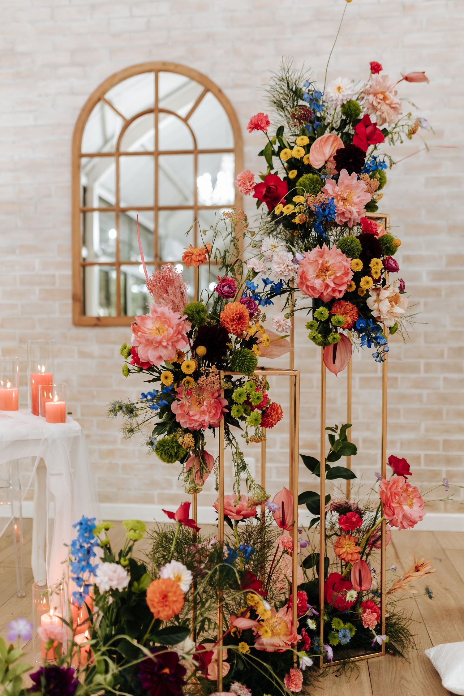 Tall floral arrangements with pink, coral and blue blooms displayed on gold stands against arched window and brick wall.