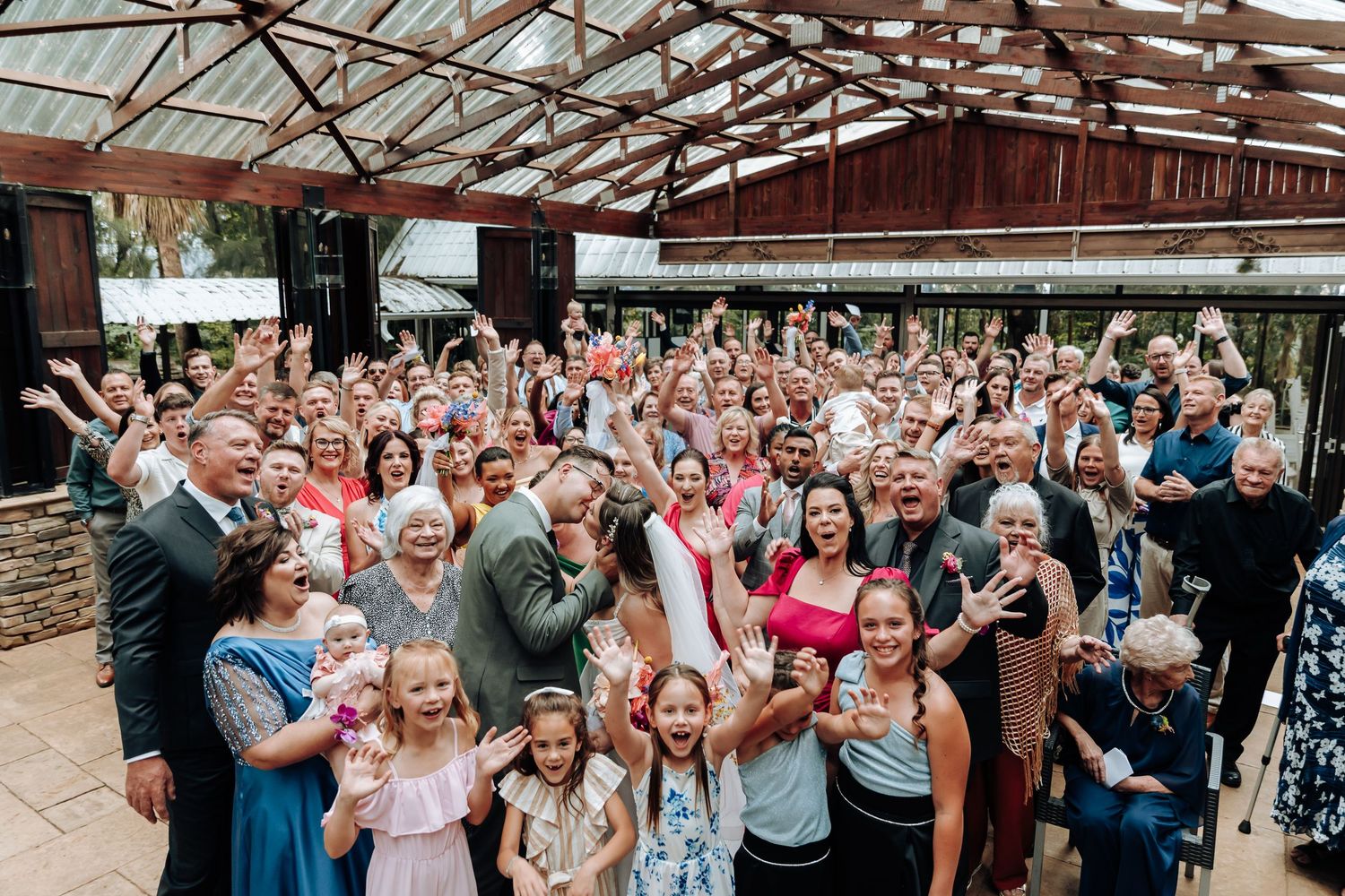 Large group wedding photo with guests gathered outdoors under rustic wooden beams.