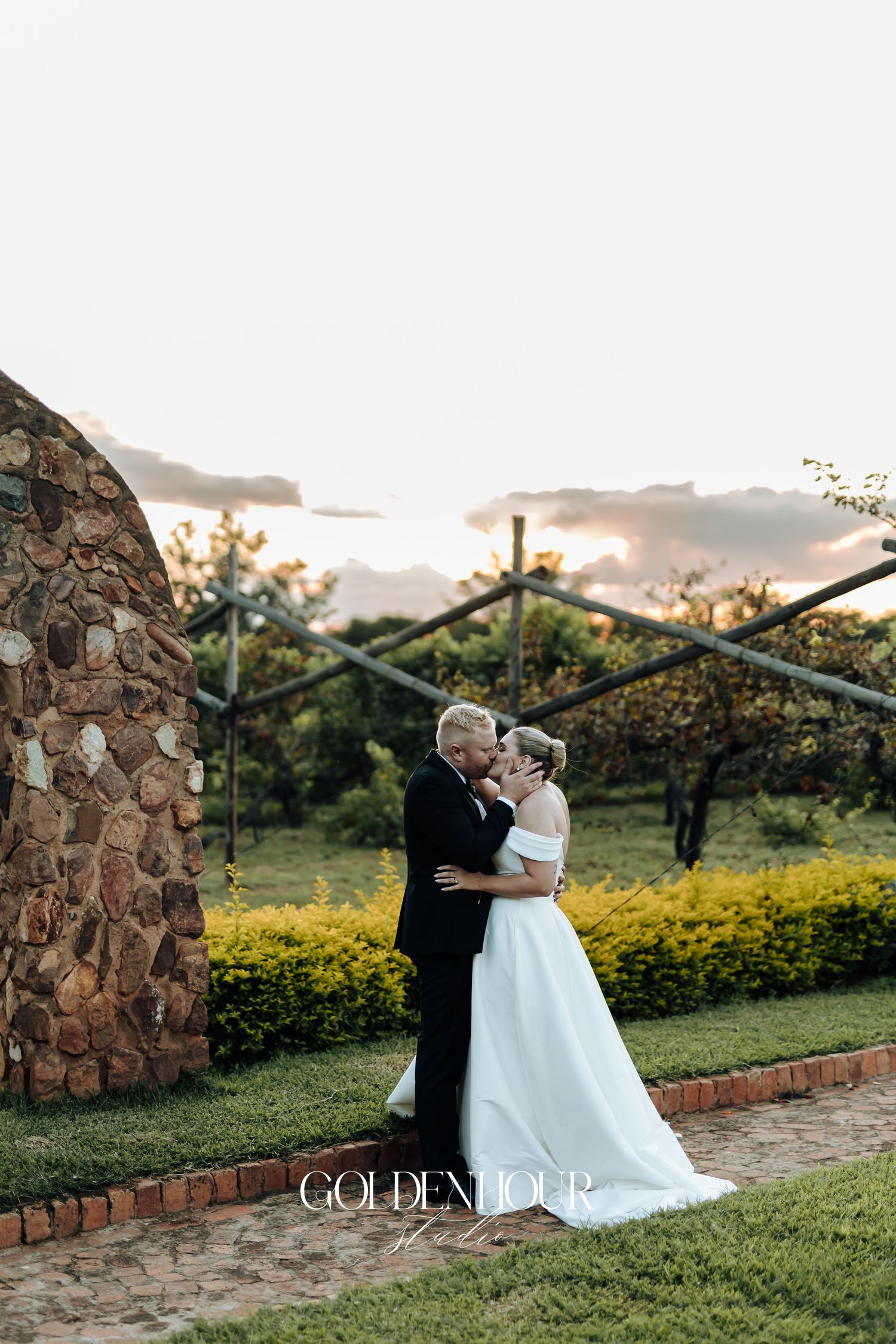 Couple in formal wedding attire sharing a romantic moment at sunset in a lush garden setting with stone architecture.