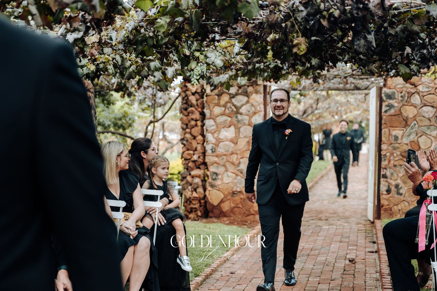 A couple walks down a brick pathway lined with autumn foliage and stone walls during an outdoor ceremony.