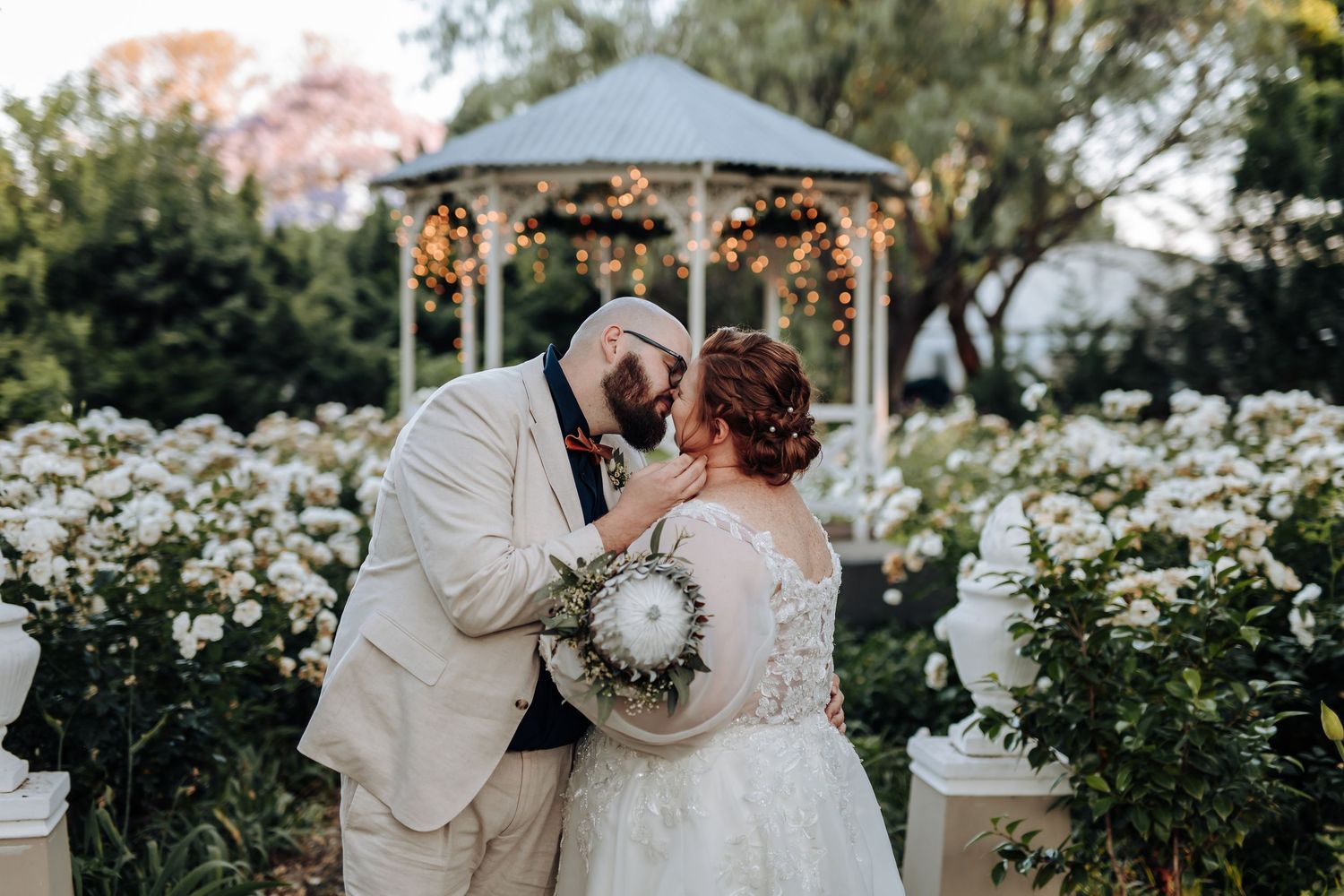 Romantic wedding couple shares tender moments in a garden setting with white flowers and a gazebo in the background.