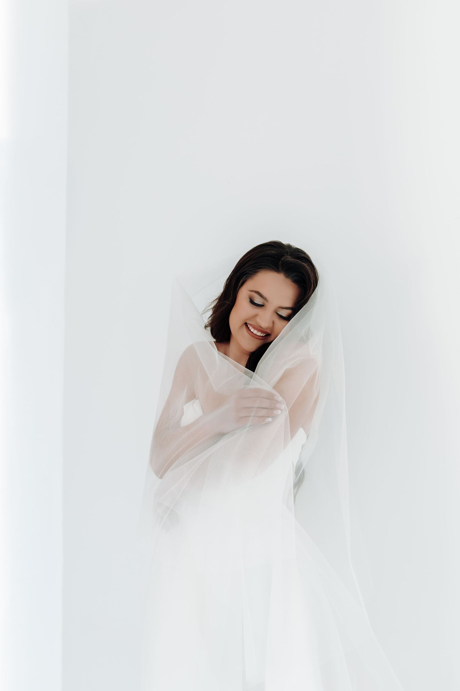 A bride in a white wedding dress and veil laughs joyfully in a bright, ethereal setting.