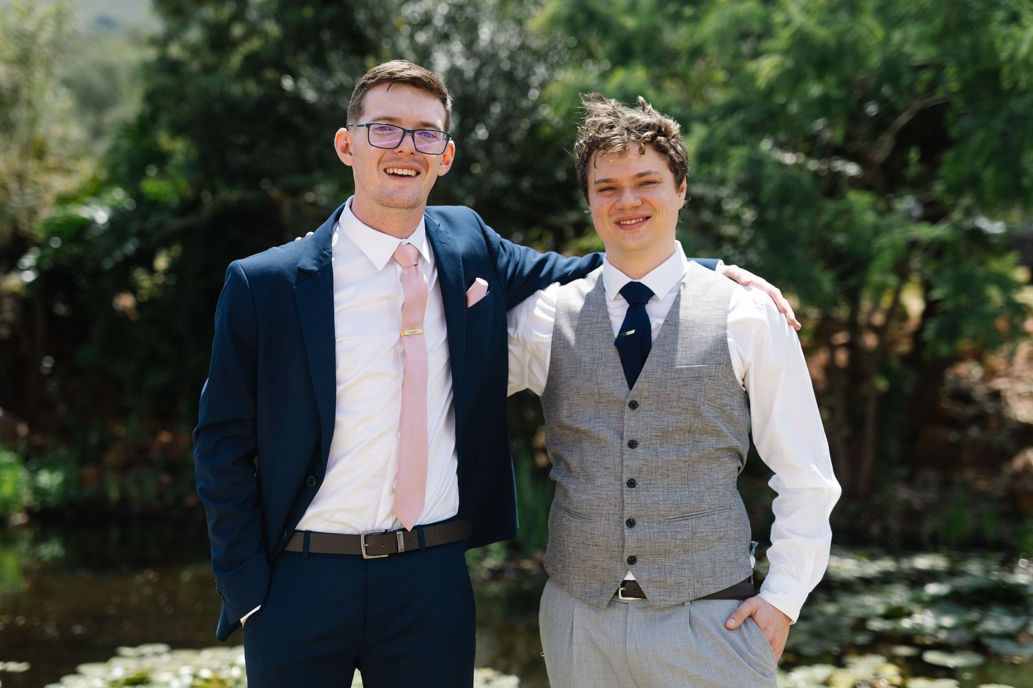 Two people in formal wedding attire stand together outdoors in a garden setting on a sunny day.