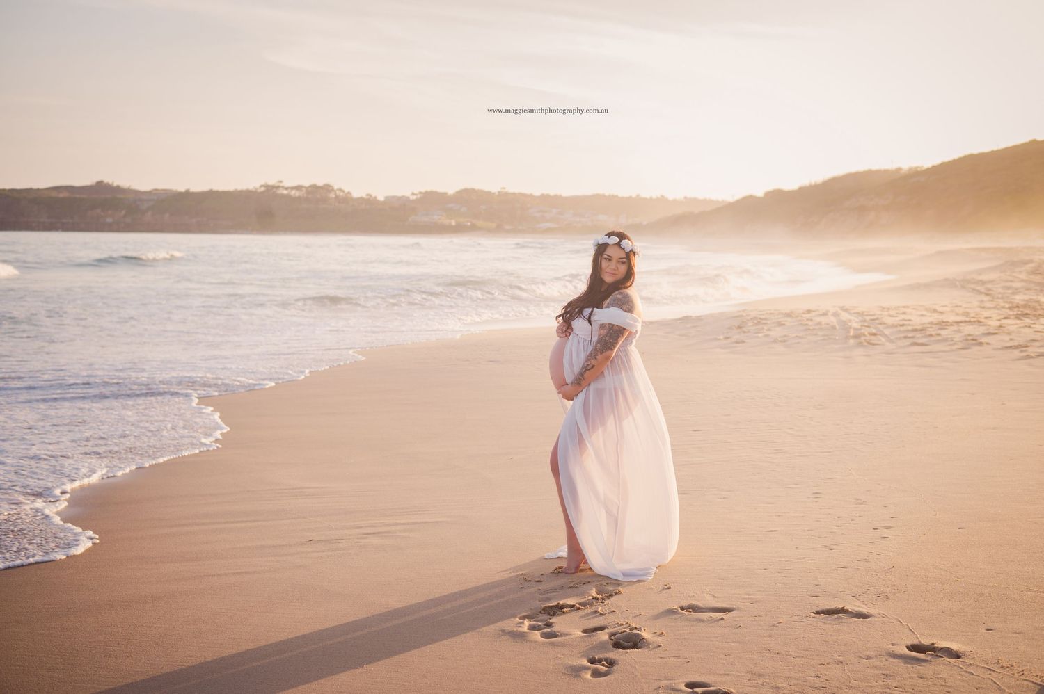 An ethereal beach scene shows a white-dressed pregnant woman on golden sand at sunset with gentle waves rolling in.