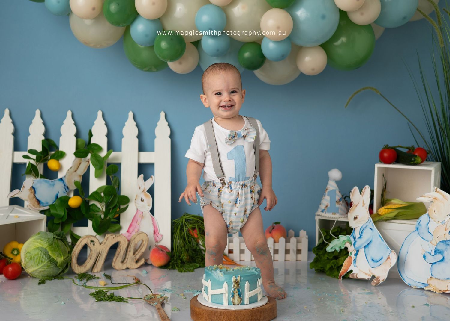 Festive cake smash birthday setup with white picket fence, green and cream balloons, and party decorations. Peter Rabbit theme