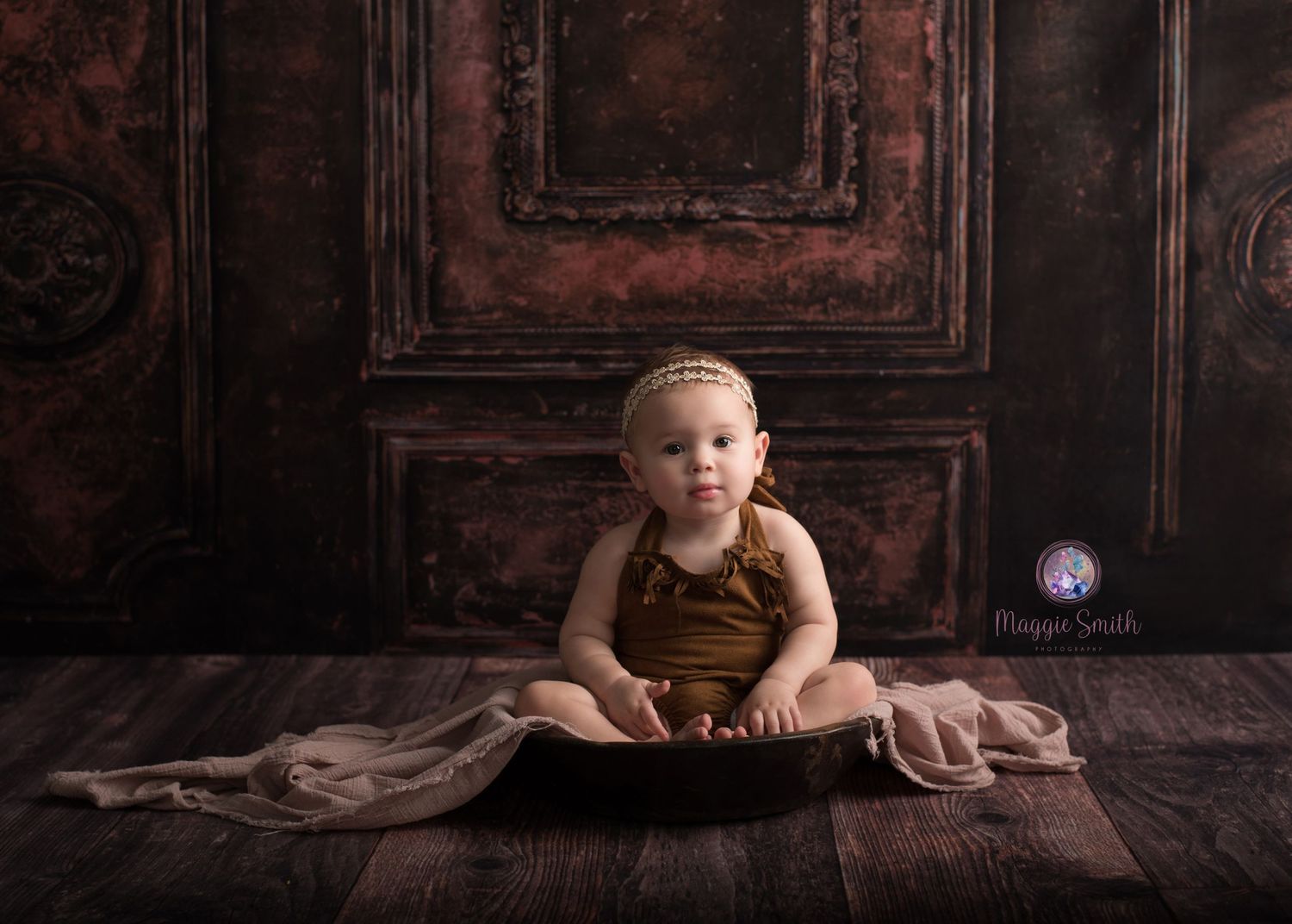 Baby wearing brown overalls sits on rustic wooden floor against vintage dark red door backdrop in studio setting.