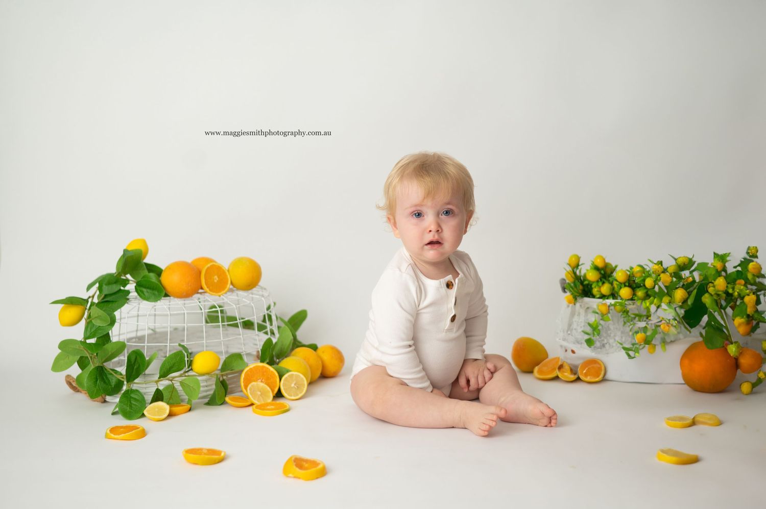A baby sits on white backdrop surrounded by fresh citrus fruits and greenery in a bright studio setting. Cake smash photography