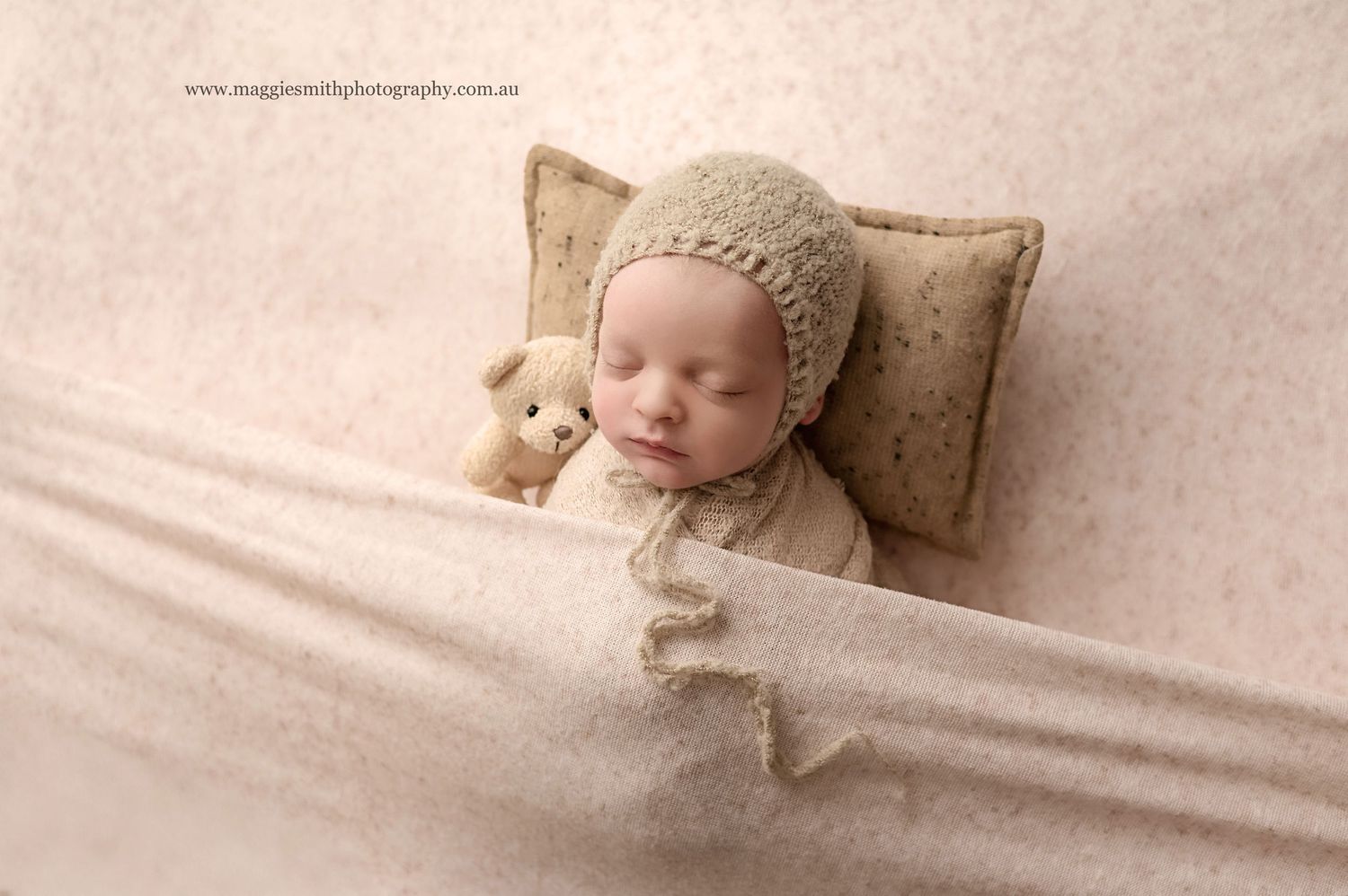 Sleeping baby wearing knit bonnet lies on brown pillow with small plush teddy bear on cream colored blanket.