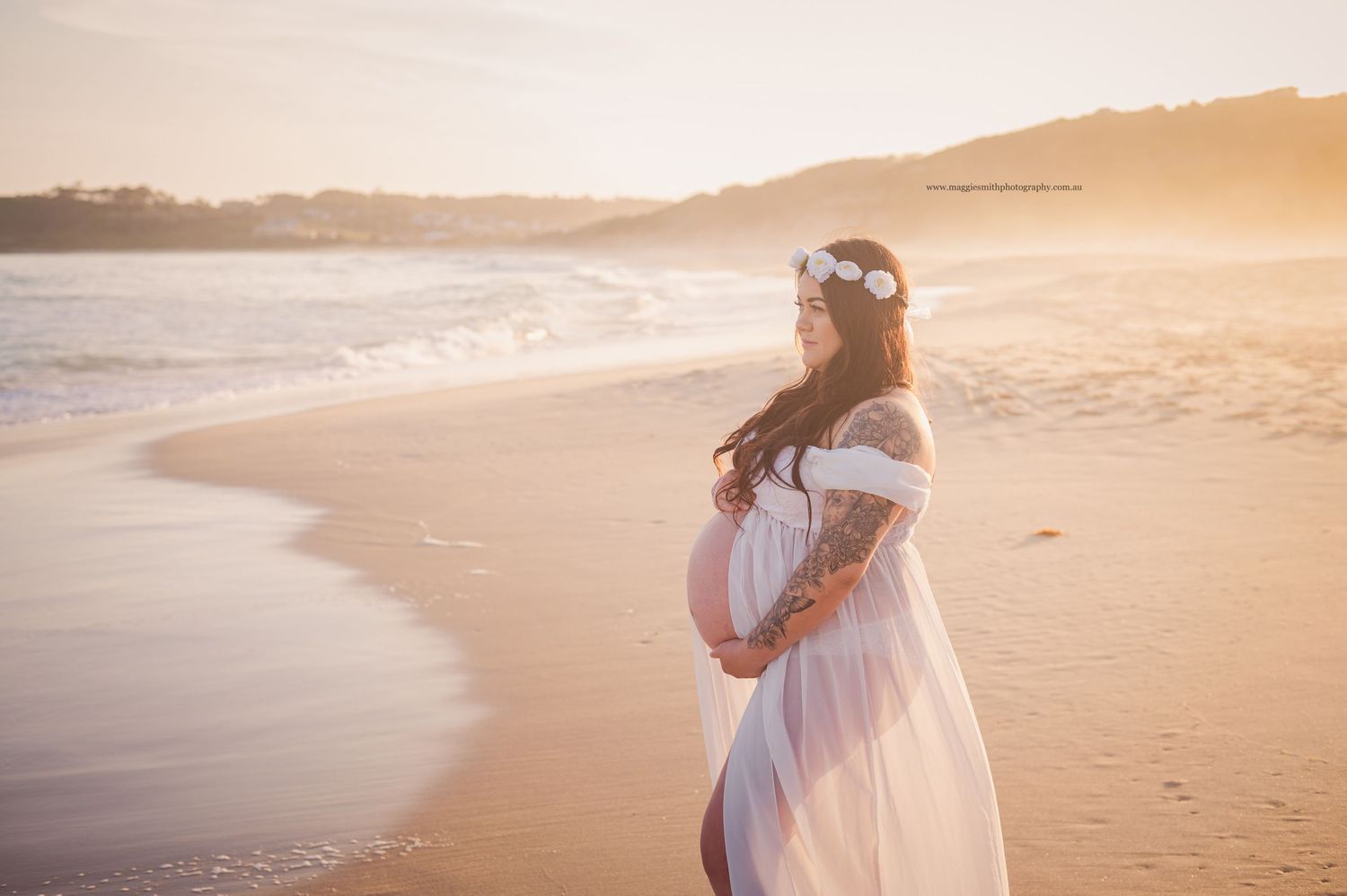 A pregnant woman in a flowing white dress stands on a beach at sunset, cradling her baby bump.