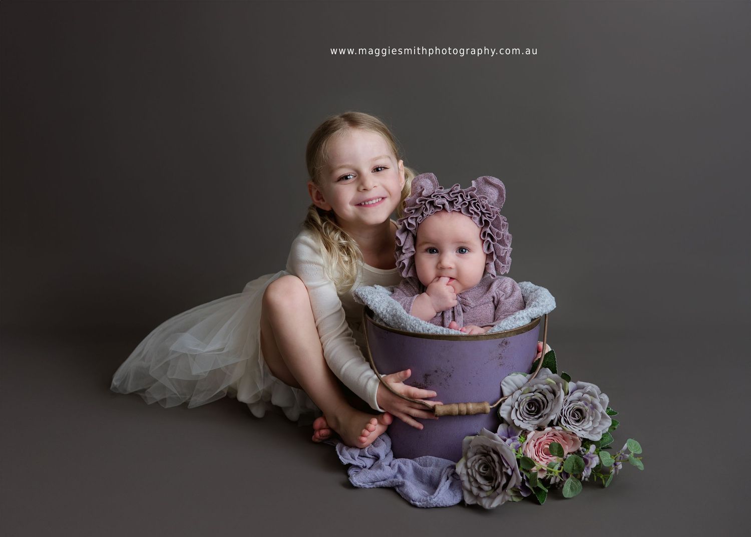 Sweet siblings pose together with lavender tub and floral decor against dark gray backdrop in studio portrait.