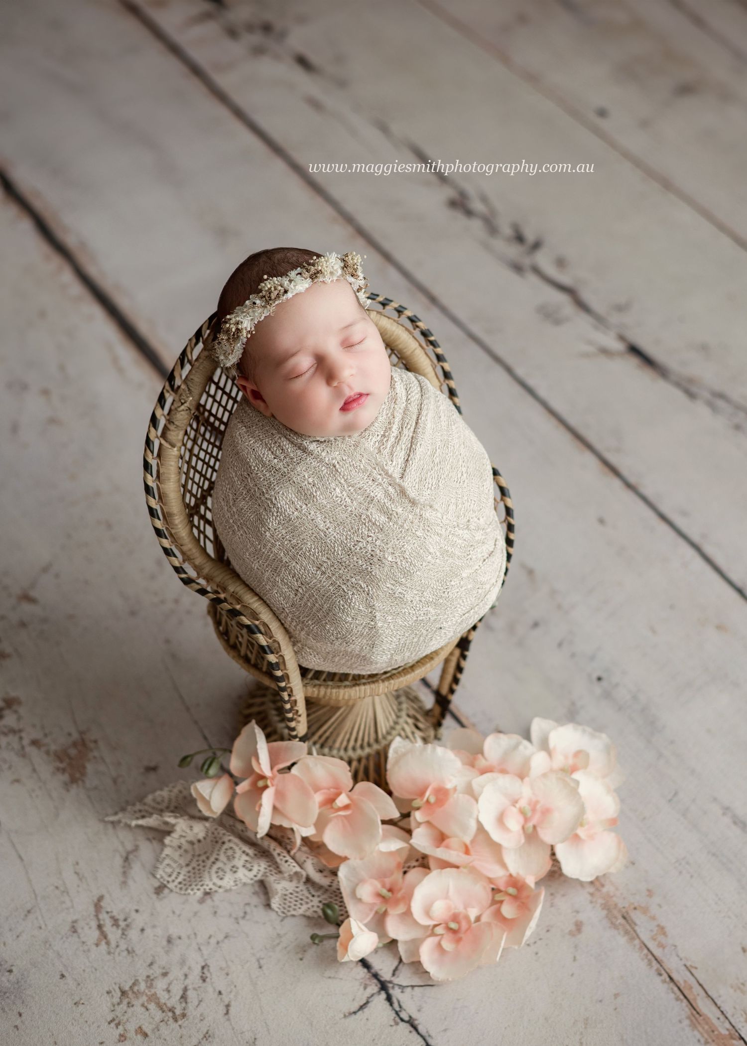 Newborn wrapped in beige fabric sleeps peacefully in wicker chair with soft pink flowers on wooden floor.