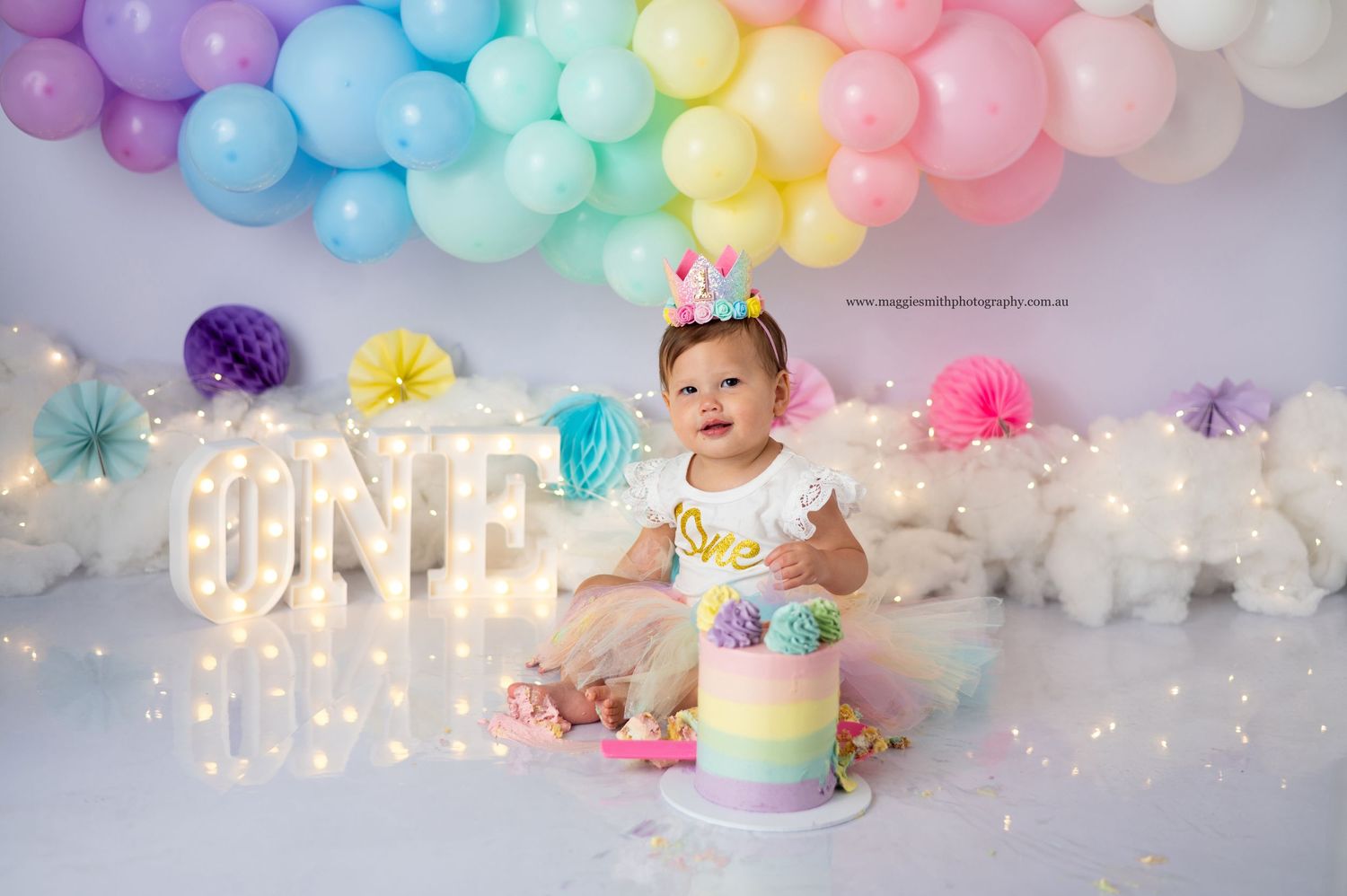 A joyful baby in colorful outfit sits by rainbow birthday cake under pastel balloon arch. Cake smash photography