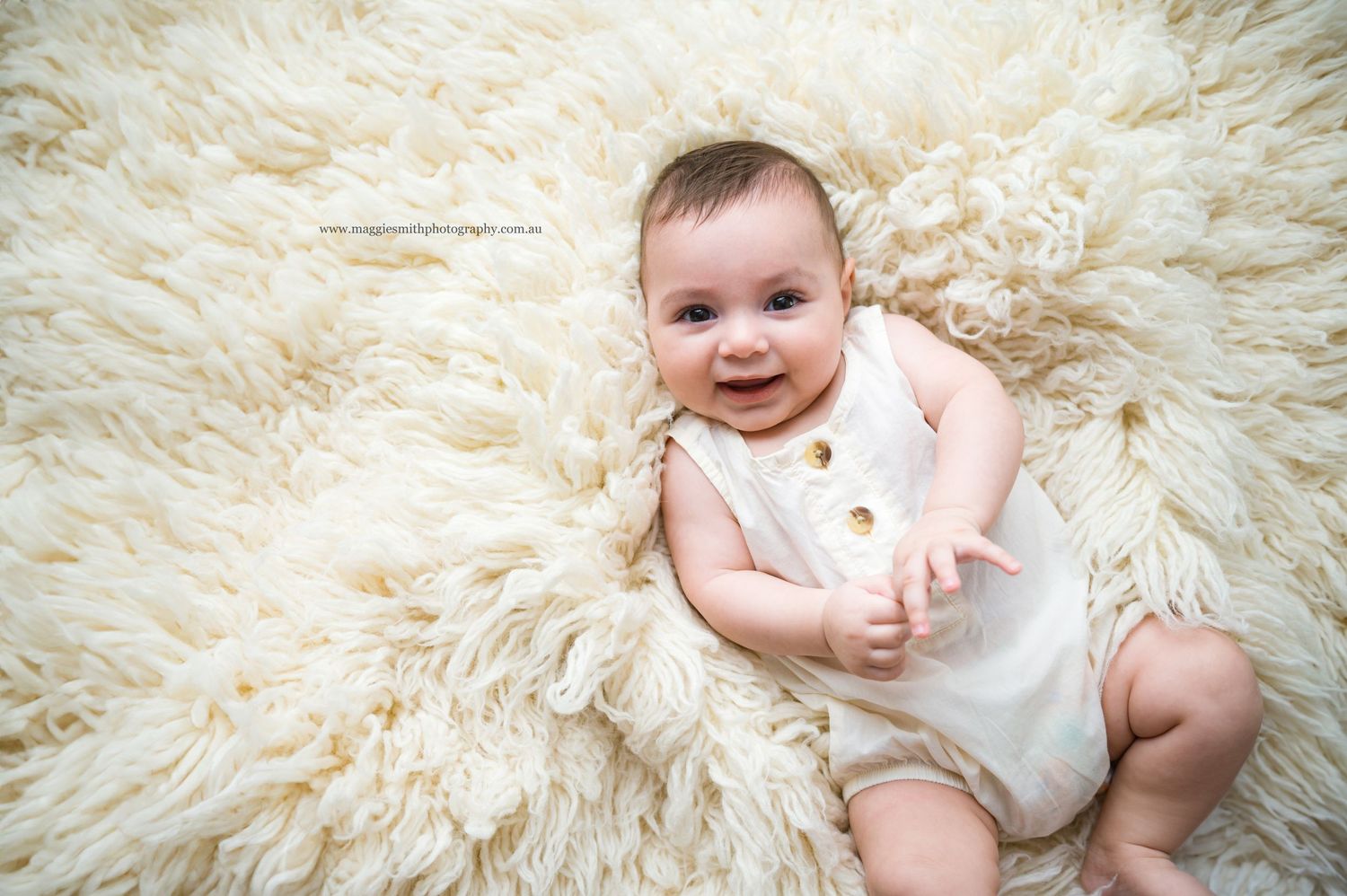 Happy baby in white romper laying on plush cream-colored fur rug during professional photo session.