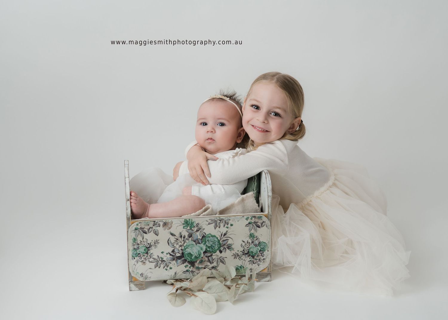 Beautiful portrait of two young children in white dresses posing with a vintage floral box against light background.