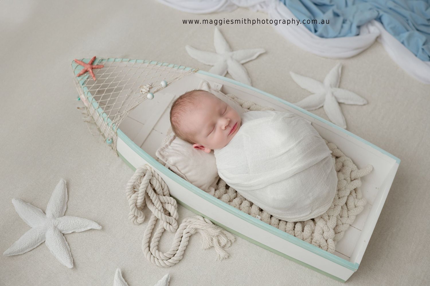 A newborn wrapped in white swaddle sleeps peacefully in a decorative boat prop surrounded by starfish decorations.