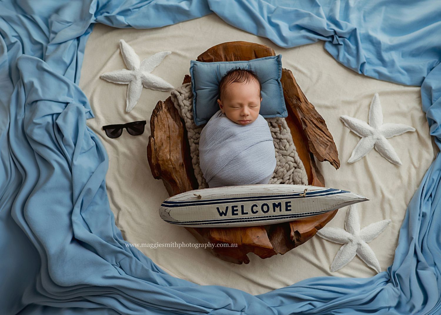 Newborn baby swaddled in blue blanket lying in wooden bowl with nautical decor and welcome sign.
