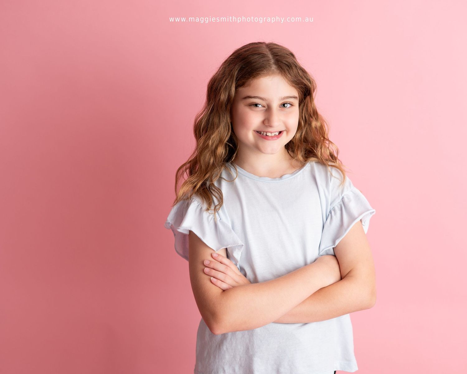 A young person with wavy brown hair wearing a light gray t-shirt stands with arms crossed against a pink background. Headshot photography