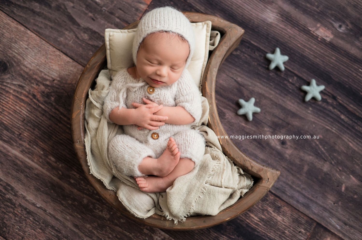 Sleeping newborn in cream knit outfit lying in wooden bowl with turquoise star decorations on dark wood floor.
