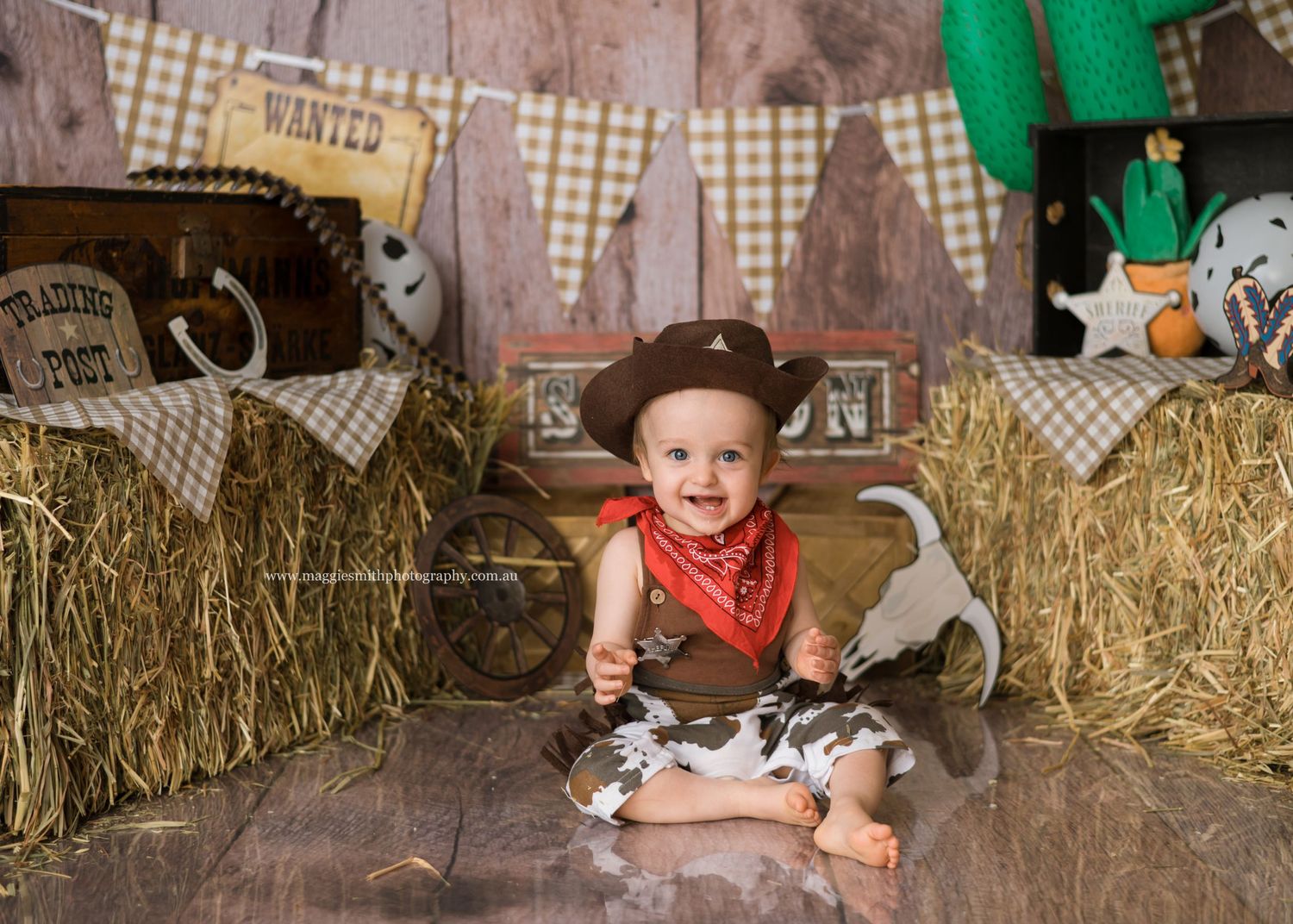 Western themed photo setup with hay bales, checkered bunting, and a small subject wearing a red bandana and cowboy boots. Cake smash photo