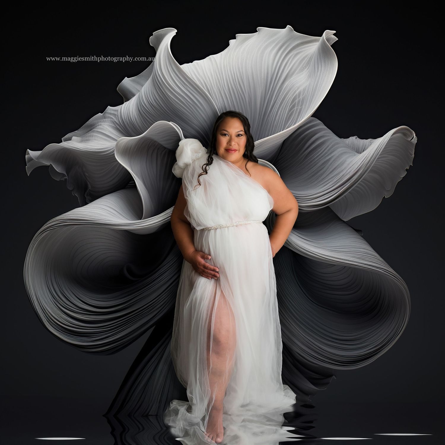 Artistic maternity portrait with flowing white dress against dramatic black backdrop featuring ethereal floral shapes.