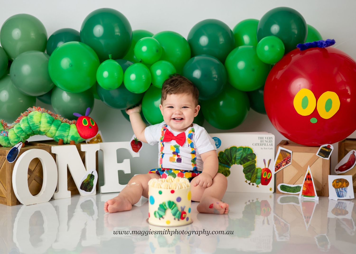 First birthday setup with green balloon arch, red caterpillar decor and white ONE letters against white backdrop. Cake smash photography.
