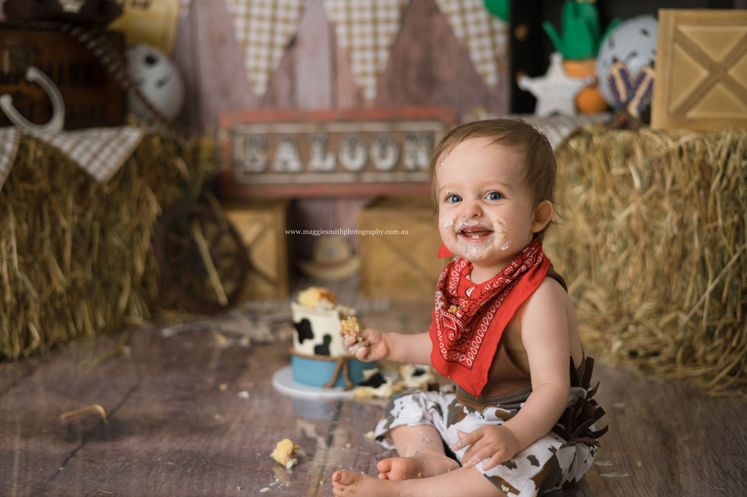 Baby wearing red bandana and denim sits among hay bales in rustic western saloon-themed setting. My first rodeo cake smash photography