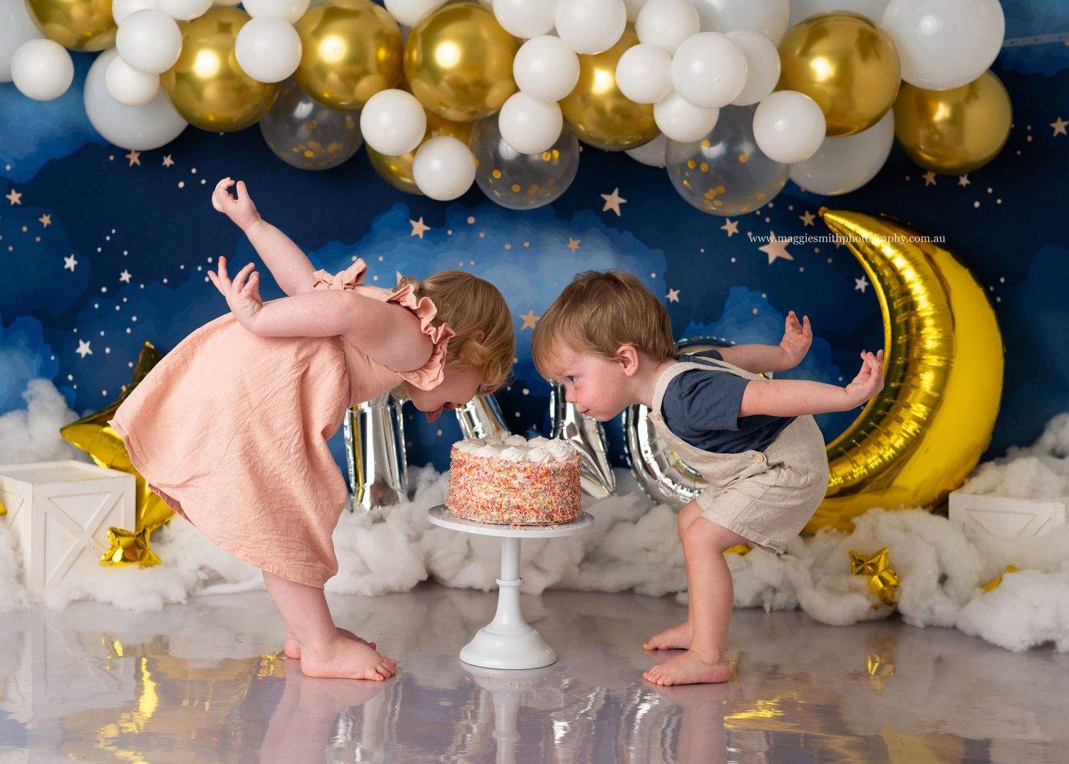 Twin boy and girl in pink and blue outfits lean forward to blow out birthday cake candles under gold and white balloon decor.