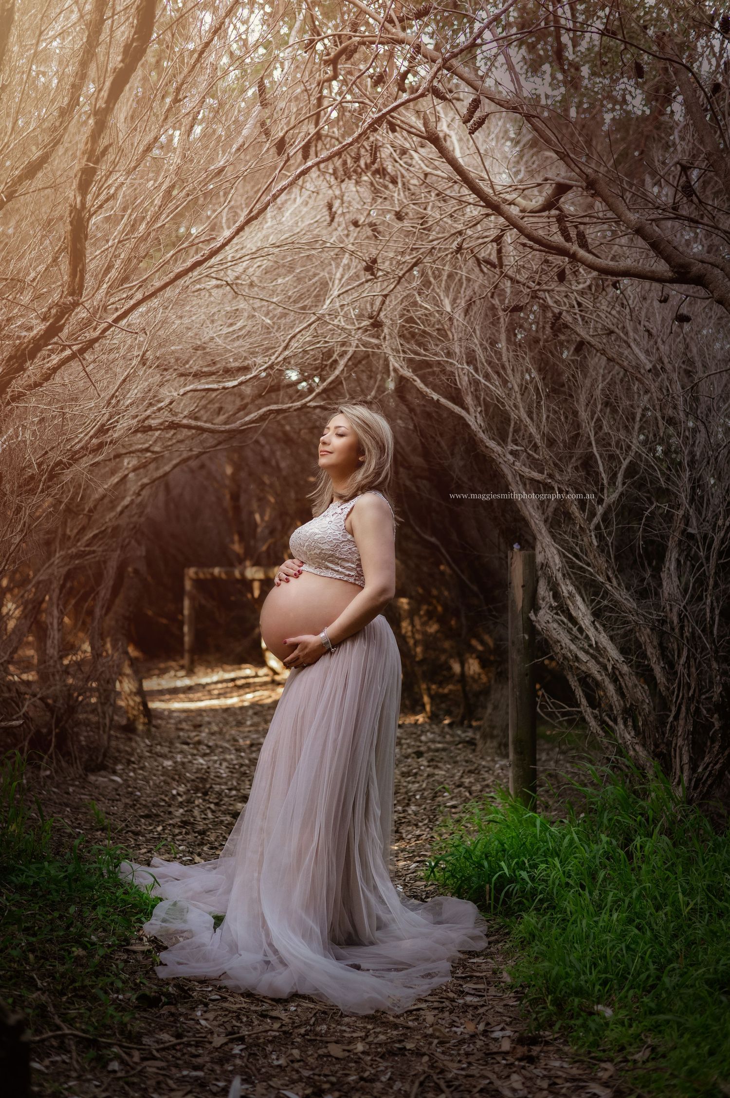 A pregnant woman in a flowing mauve dress stands on a woodland path as golden sunlight filters through the trees.