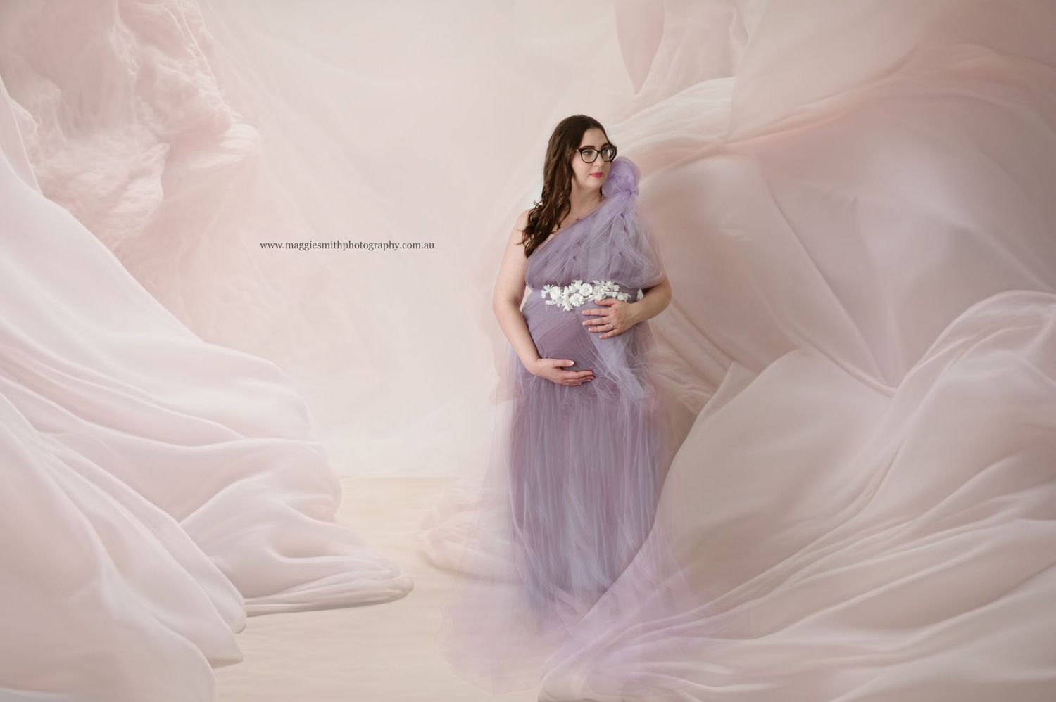 Ethereal maternity portrait with flowing lavender tulle fabric creating dreamy atmosphere against soft pink background.