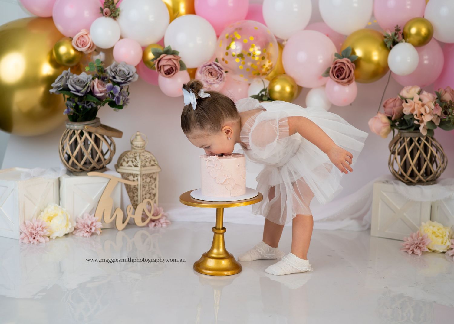 A festive party setup with pink and gold balloon garland, lanterns, and a little girl leaning over a cake on a gold stand.