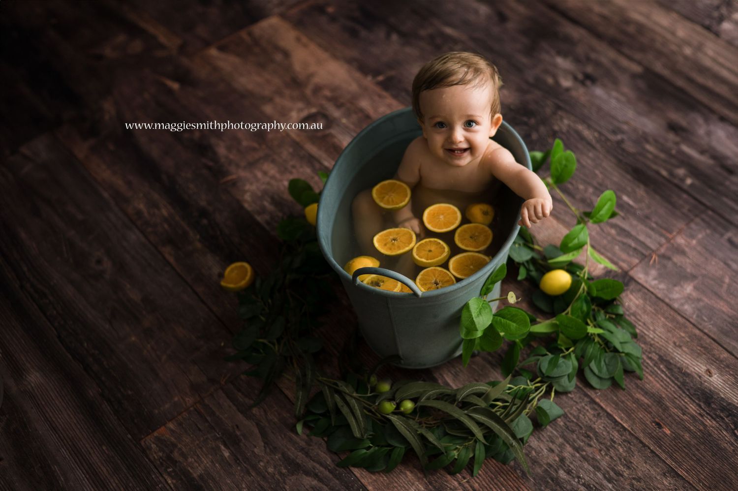Baby sitting in a teal bathtub with citrus slices and greenery on dark wooden floor during a photography session.