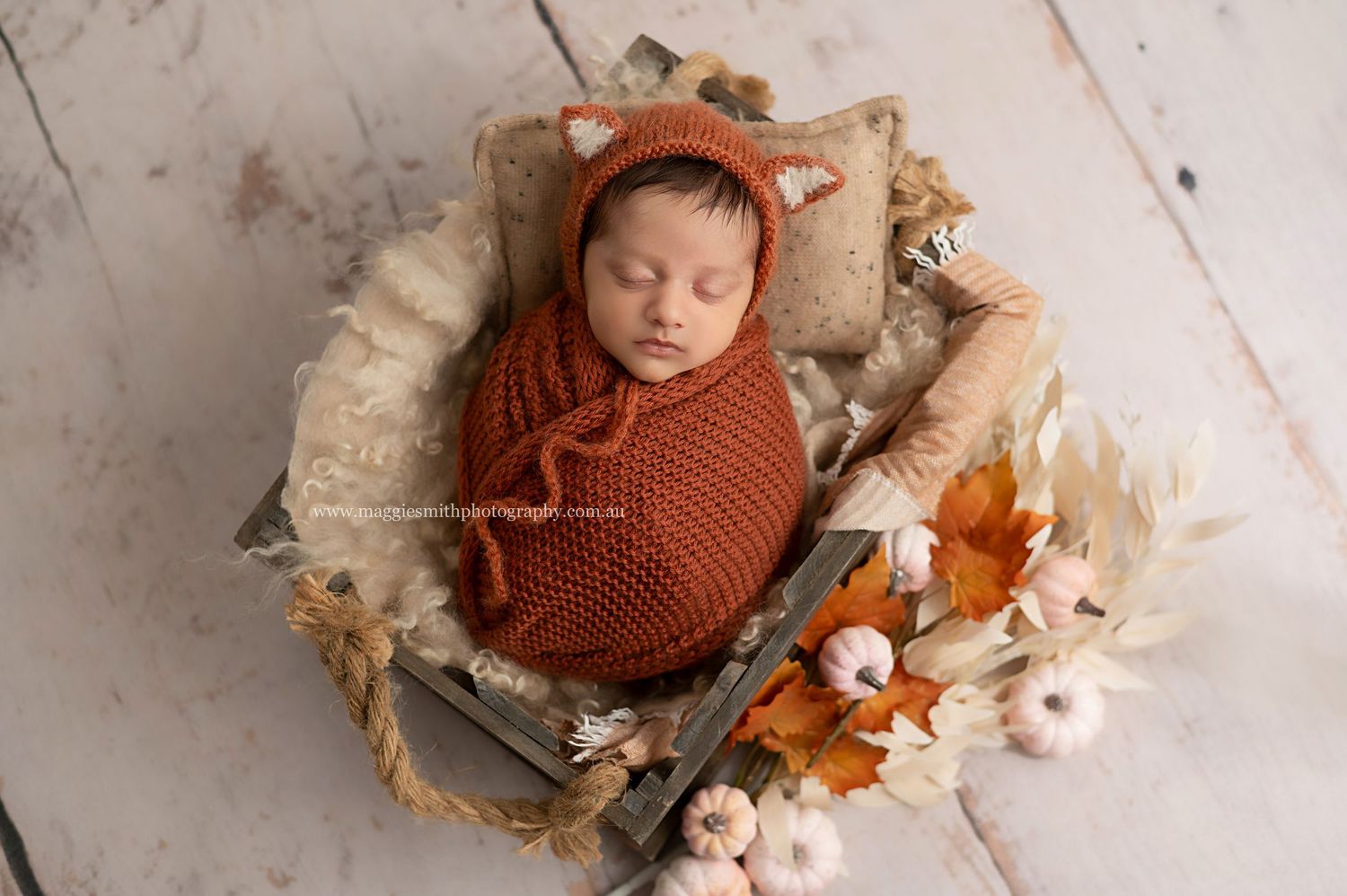 Newborn in rust-colored knit wrap sleeps peacefully in wooden basket surrounded by autumn-toned dried flowers.