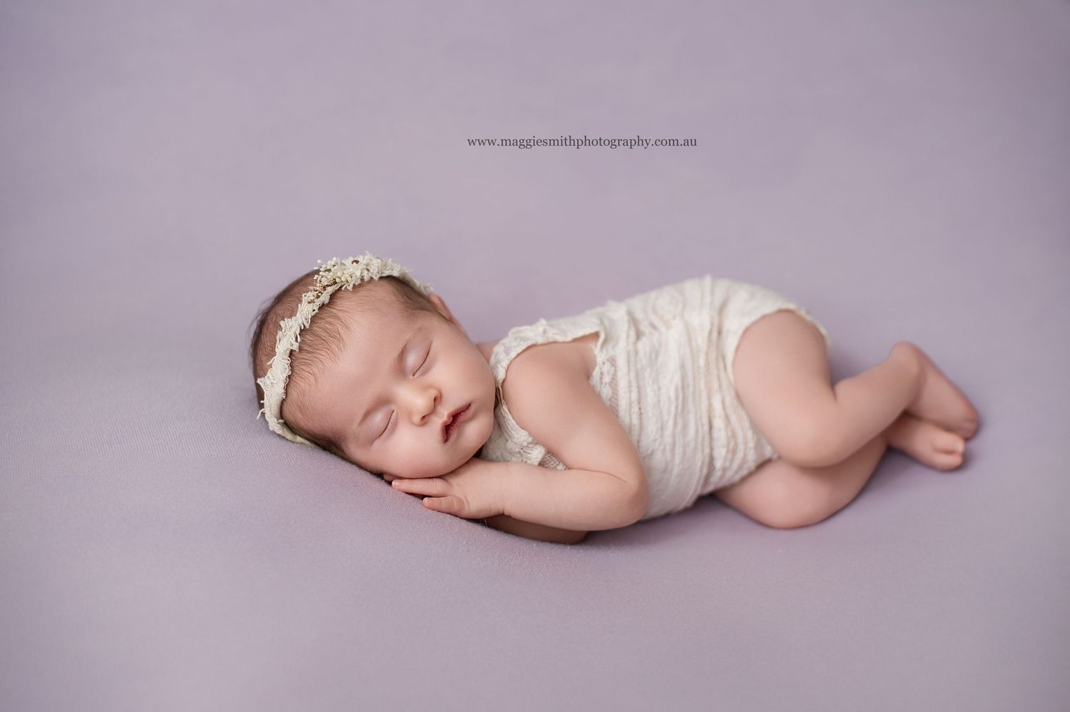 A sleeping newborn baby wearing white lace lies curled up on a soft purple blanket in a studio setting.