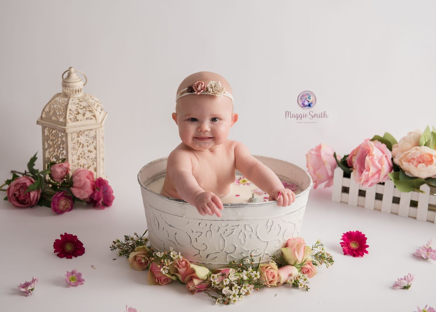 Baby sits in decorative white bowl surrounded by pink flowers and lantern for milestone photo session.
