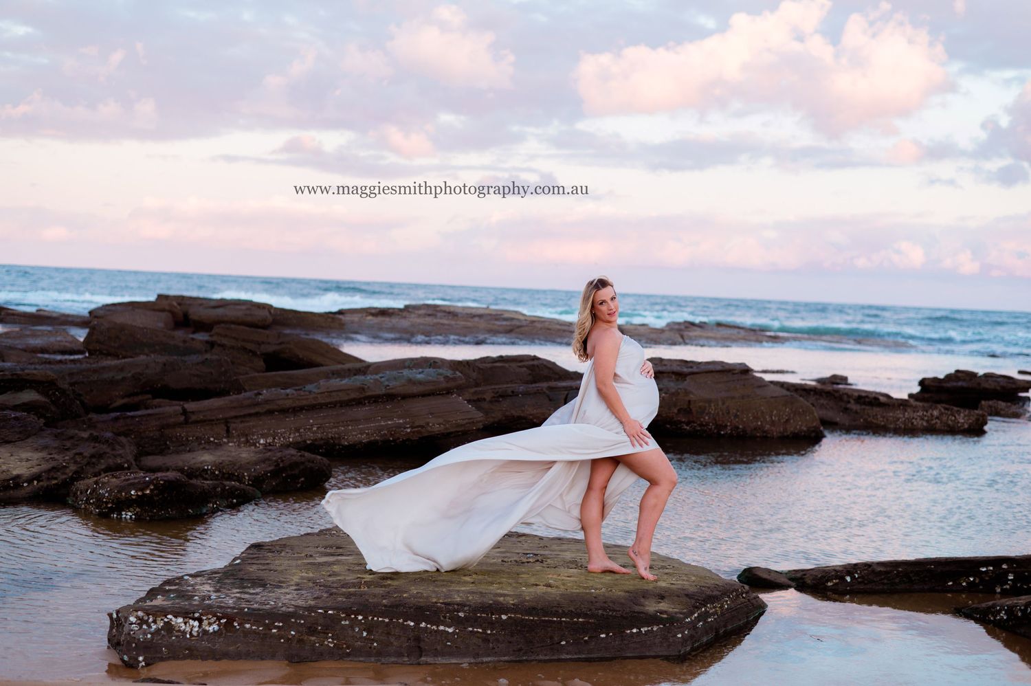 A flowing white maternity dress catches the ocean breeze as a pregnant goddess poses on coastal rocks at sunset.