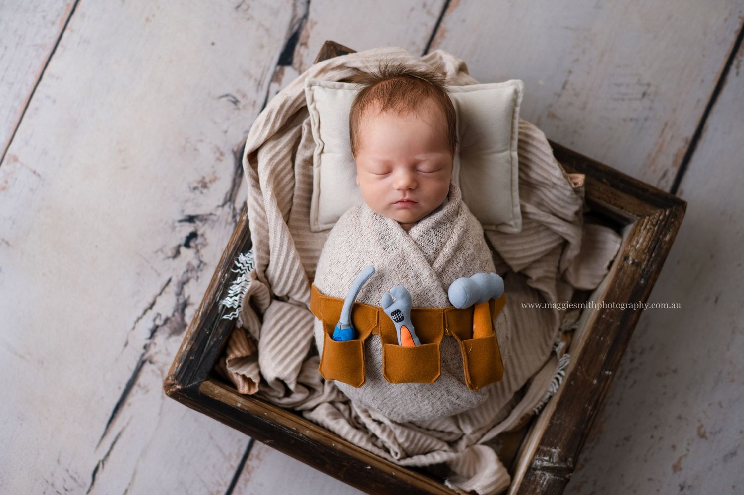 A sleeping newborn wearing a tool belt rests peacefully in a rustic wooden crate lined with cozy blankets. Newborn photography Central Coast
