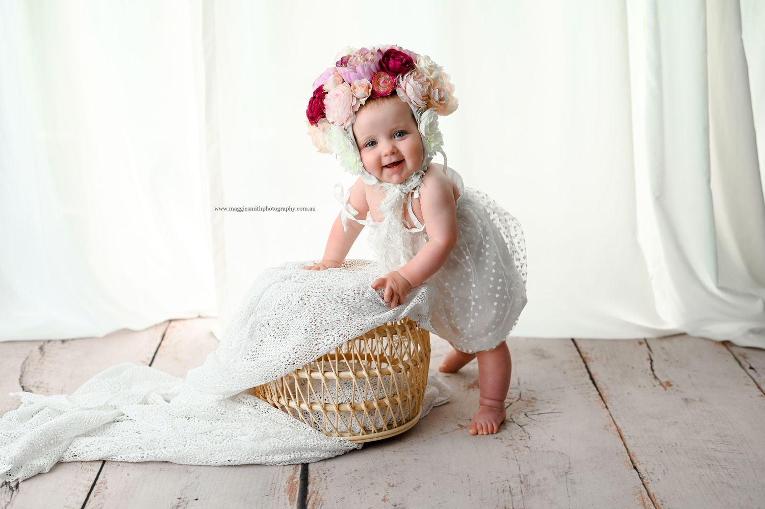 A toddler in a white dress and floral headband plays with a woven basket on wooden floorboards with white curtains behind.