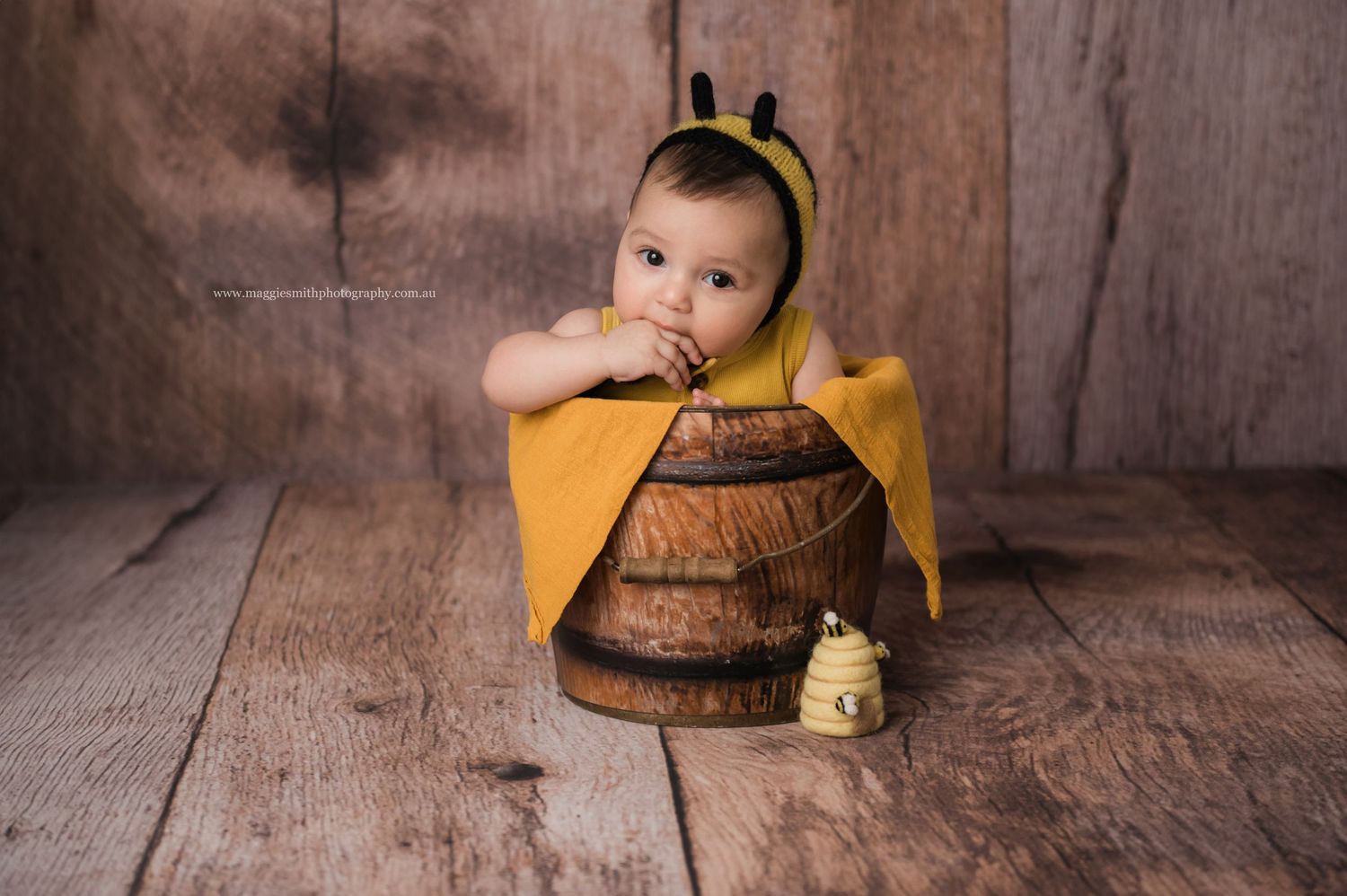 Small baby wearing yellow and black bumble bee costume sits in rustic wooden barrel on wooden floor.