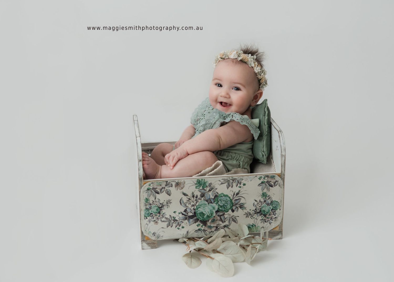 Baby wearing mint green outfit sits in vintage-style floral patterned box during professional portrait session.