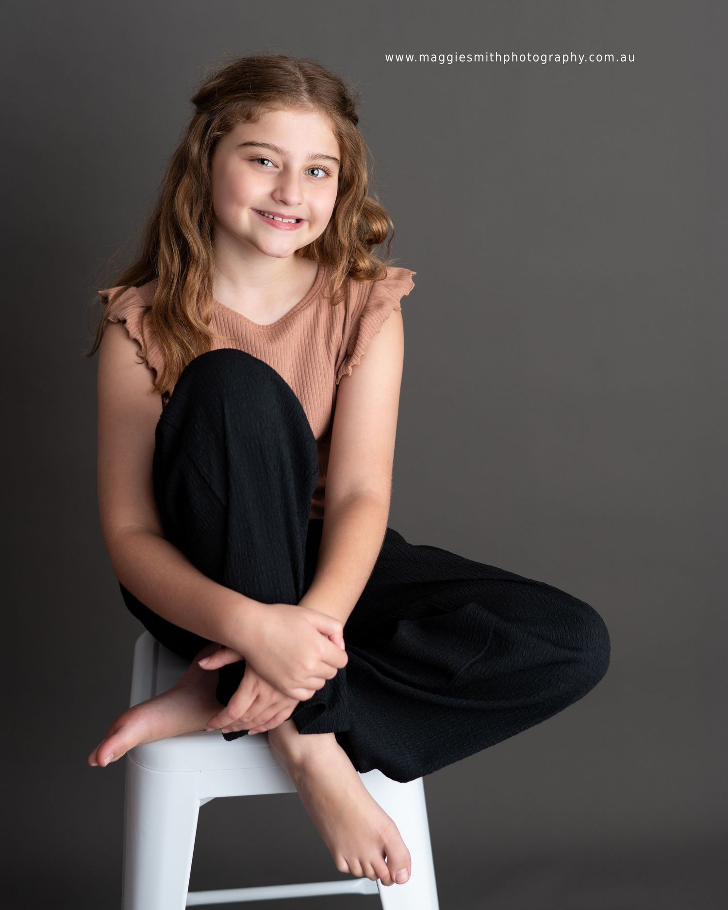 Professional portrait photography of a young person in black pants sitting casually on a white stool against a gray backdrop.