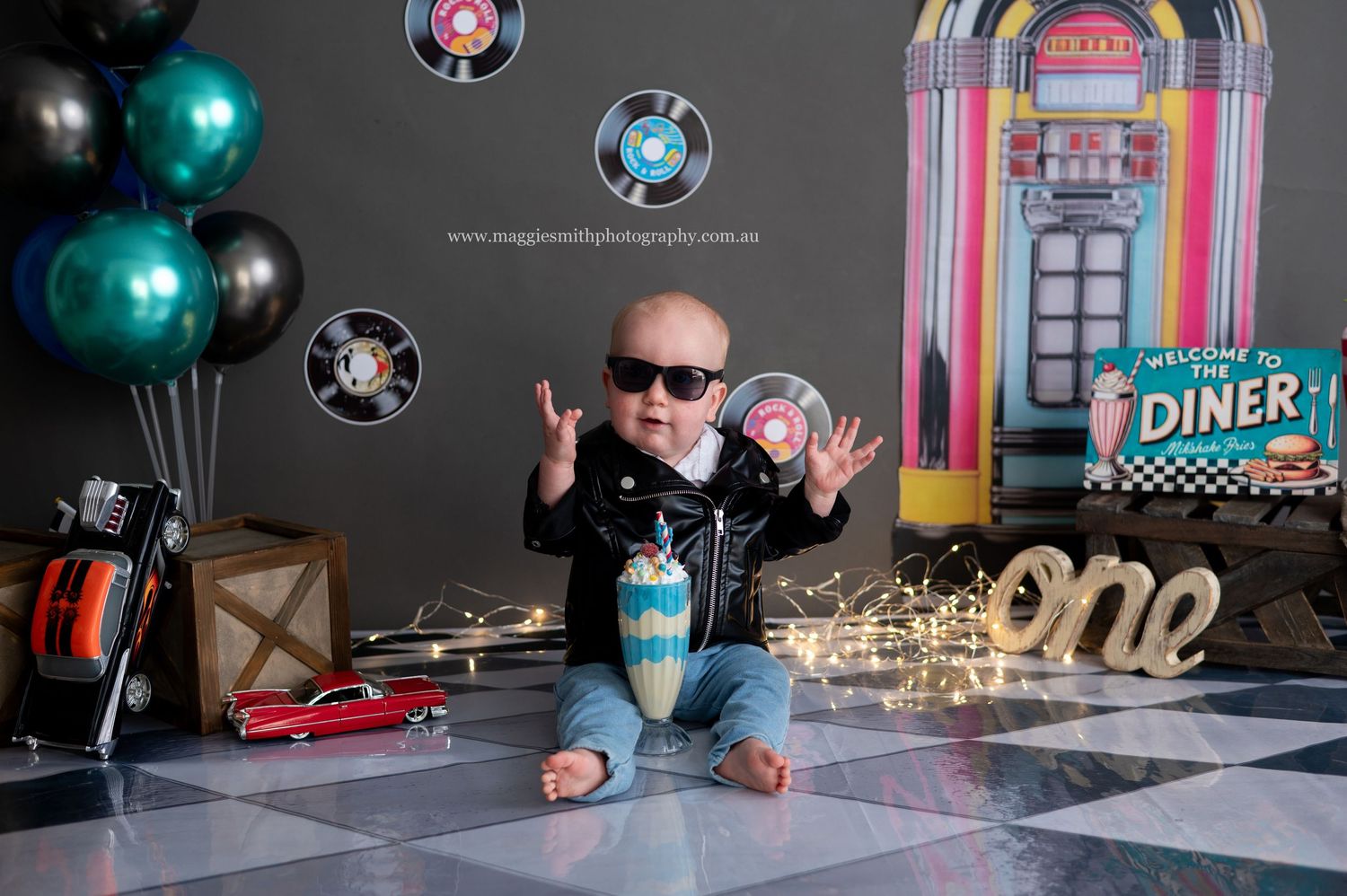 Retro diner themed party setup with balloons, neon signs, and milkshake decorations in black and turquoise colors. Cake smash photography
