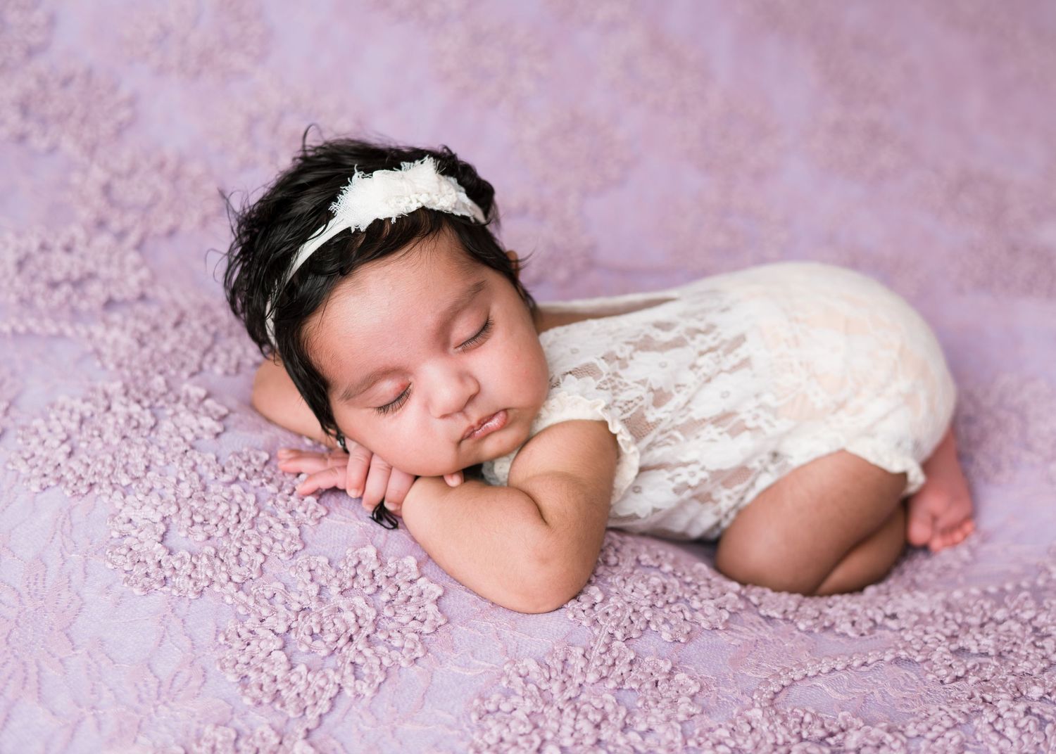 Sleeping newborn in white lace romper and headband poses peacefully on soft pink textured backdrop.
