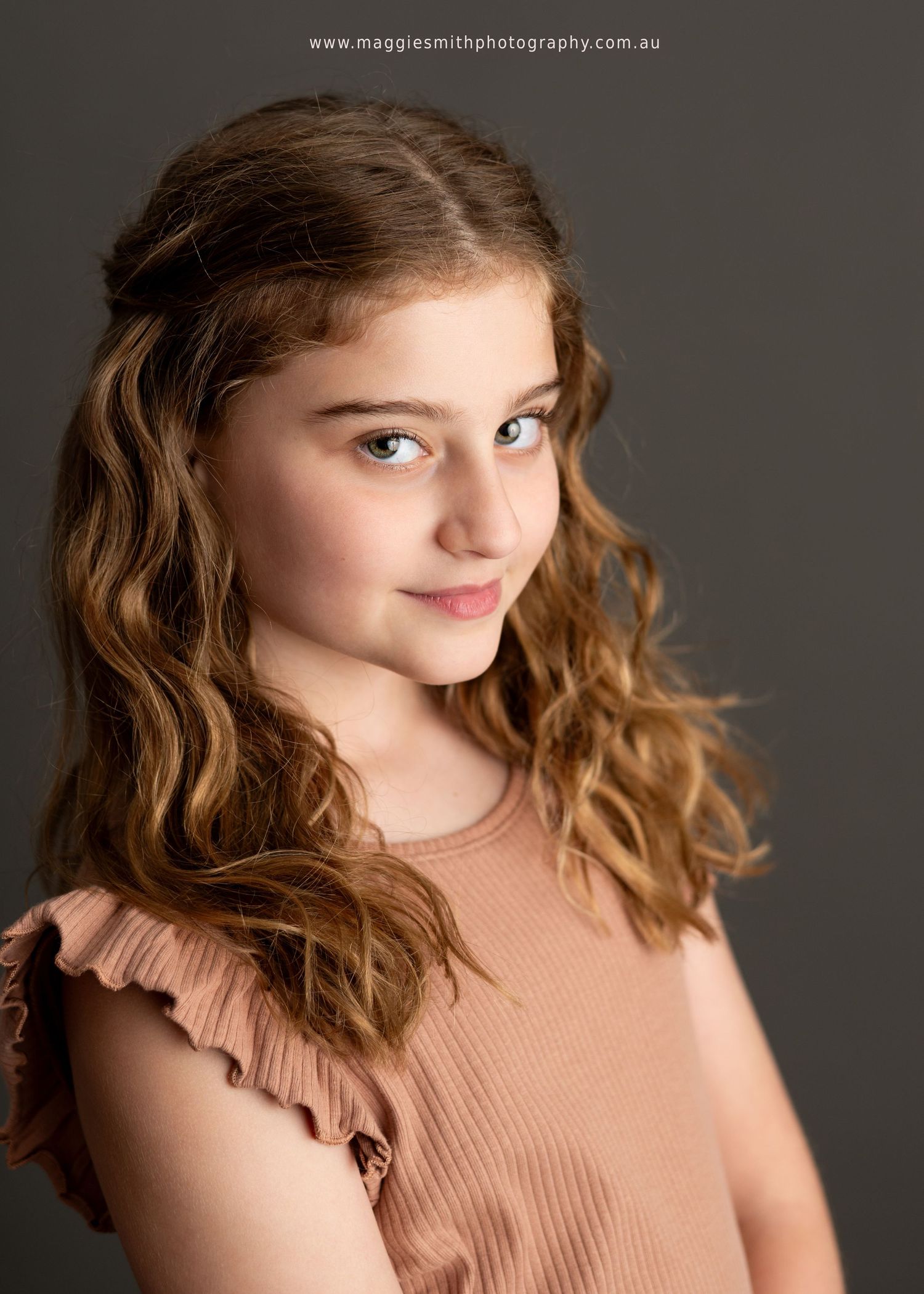 Professional portrait of young person with wavy brown hair wearing a peach-colored ruffled top against dark background.