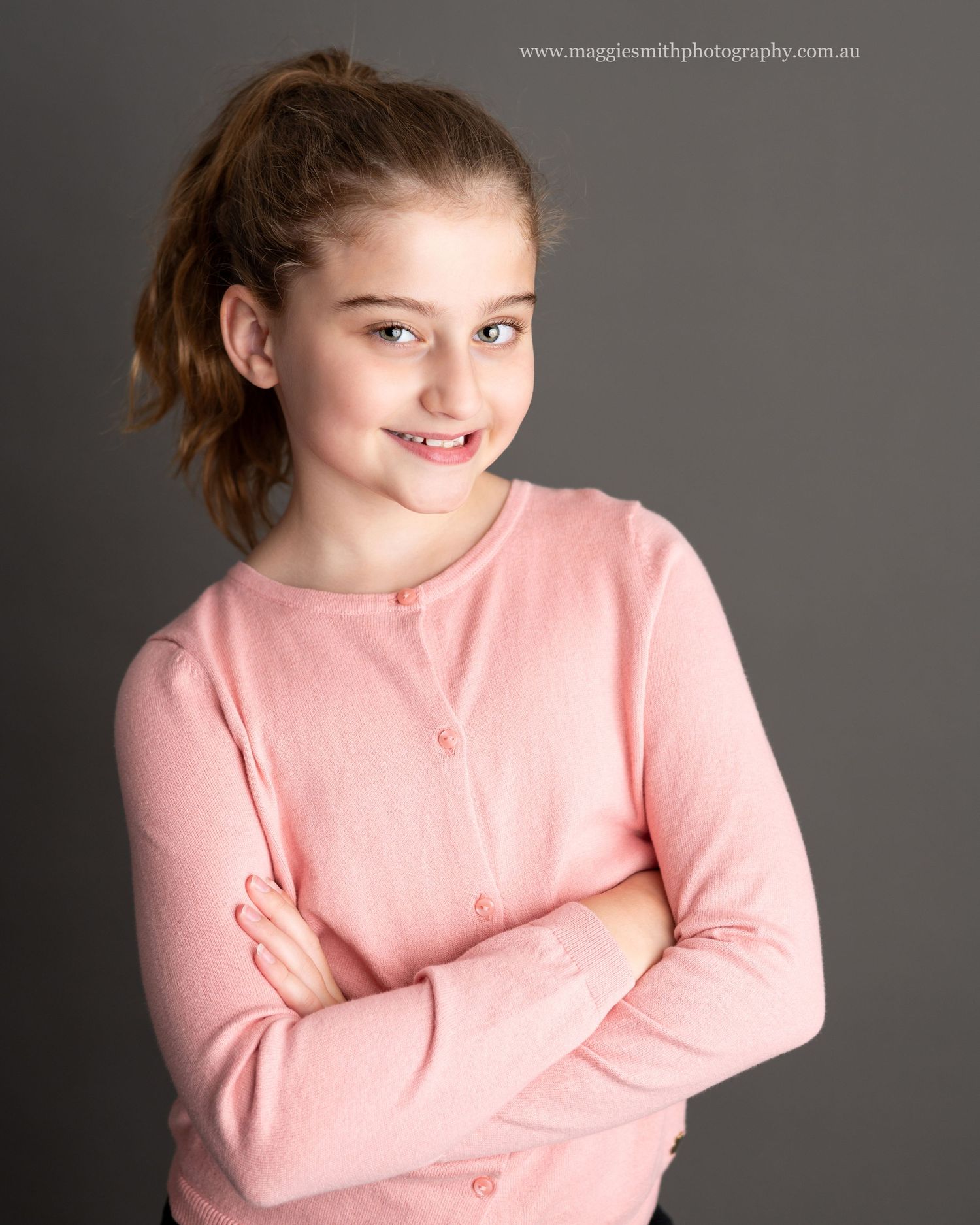 Portrait of girl in a soft pink cardigan sweater smiling warmly against a dark grey background. Headshot photography