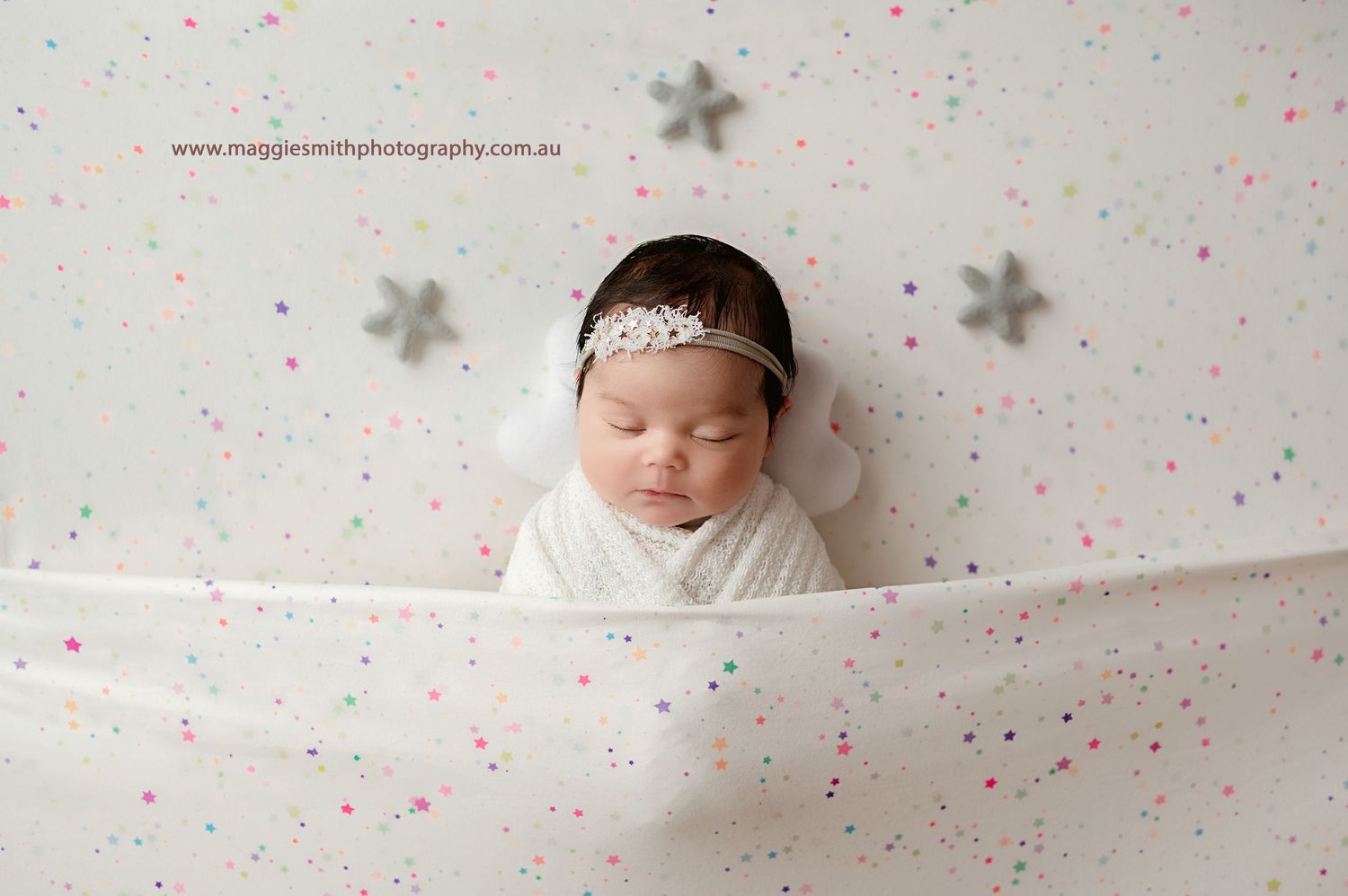 A sleeping newborn wrapped in white fabric against a backdrop with pink dots and silver stars. Central Coast newborn photography
