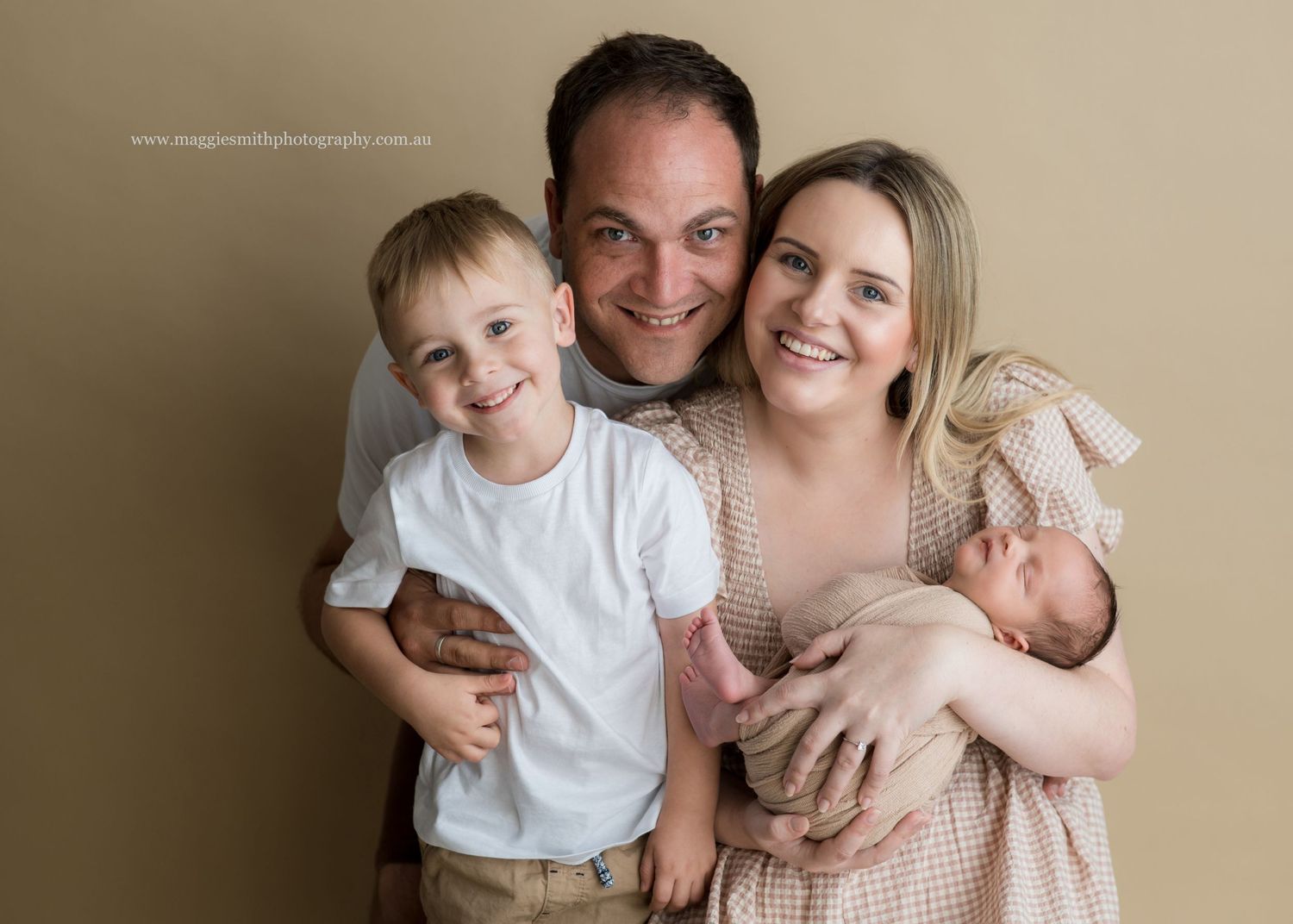 Portrait of a loving family with young child and newborn baby against a light beige backdrop.