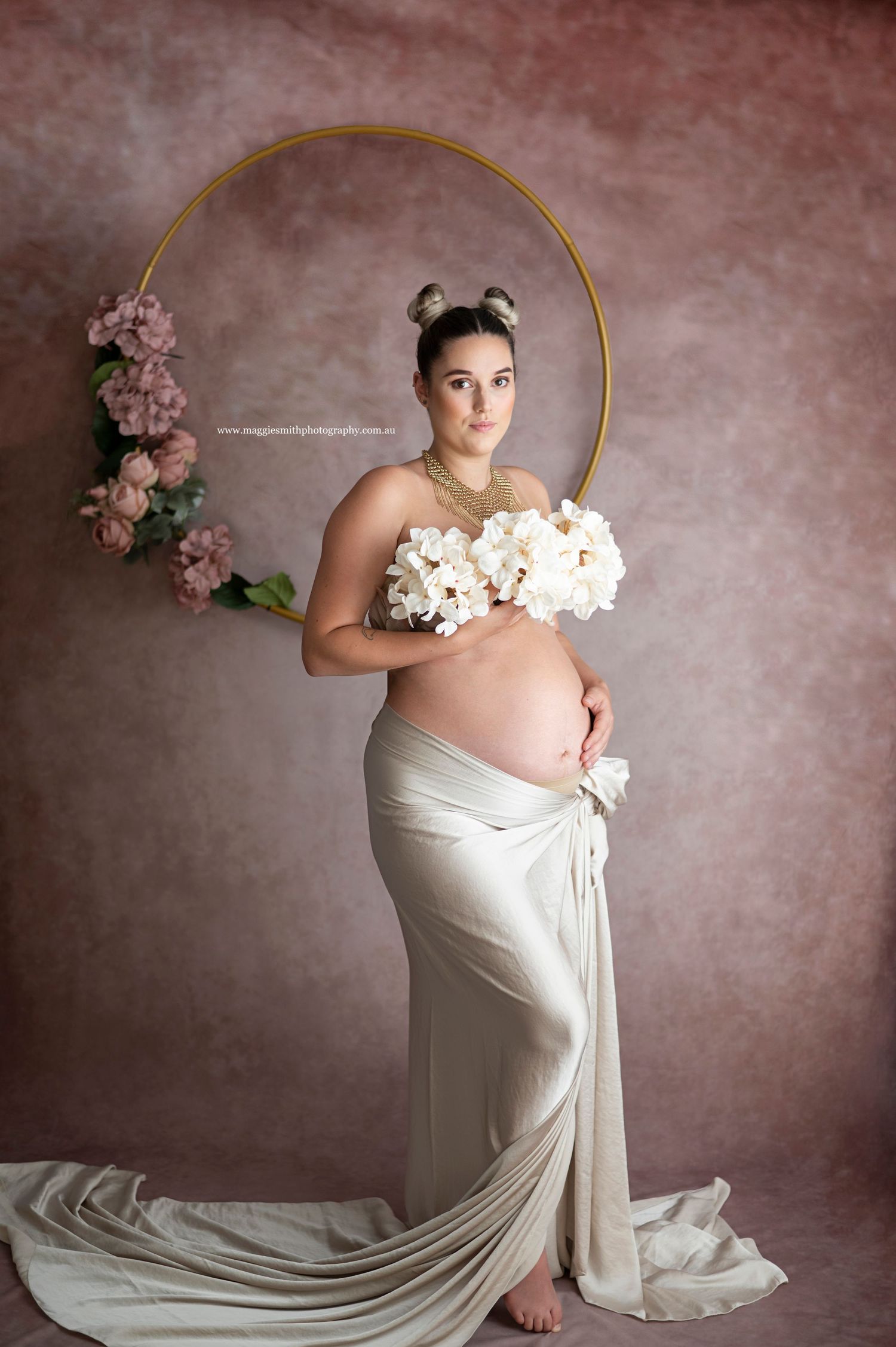 Elegant maternity photo featuring a draped white gown, golden hoop arch and pink floral accents against textured mauve backdrop.