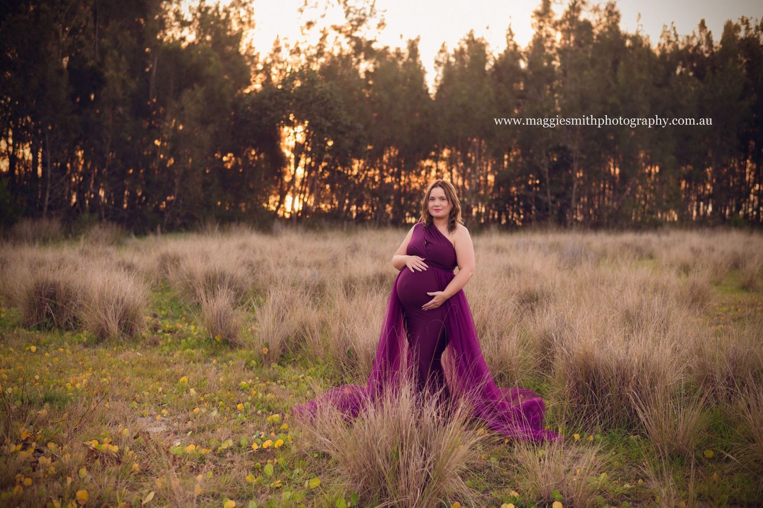 A flowing purple maternity gown billows in the breeze amid tall grass at sunset with pine trees in the background.
