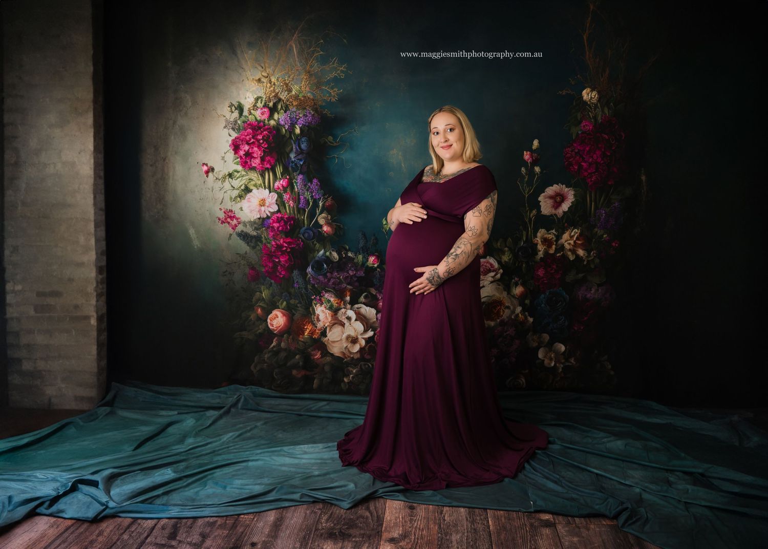 A maternity portrait in a plum colored gown against a dark backdrop with vibrant floral arrangements.