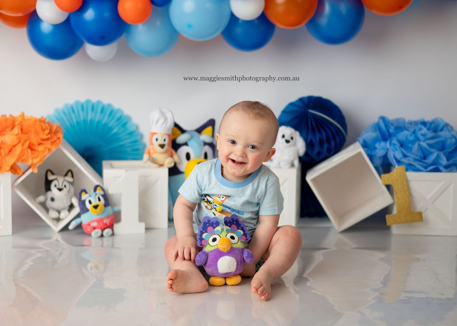 A festive birthday setup with blue and orange balloons, Bluey theme, and a happy baby sitting on the floor. Cake smash photography